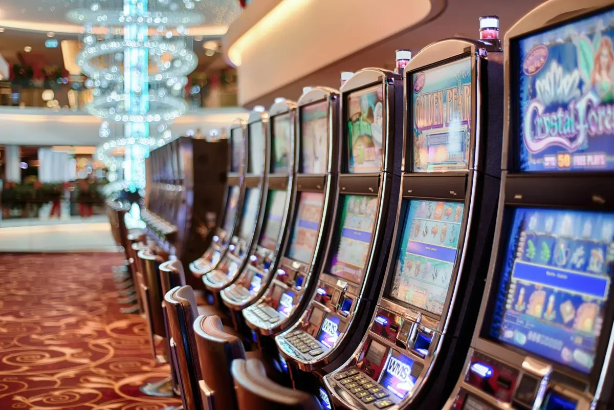 Row of modern electronic slot machines in a brightly lit casino with a decorative chandelier in the background