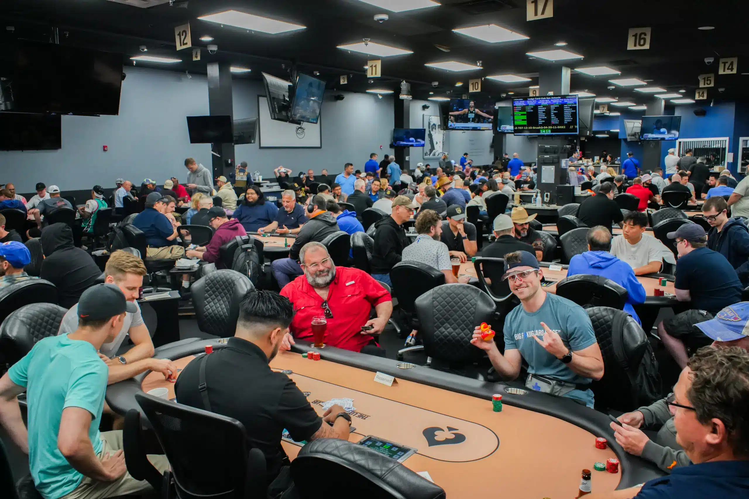 A busy poker tournament card room with many players at tables and monitors in view.