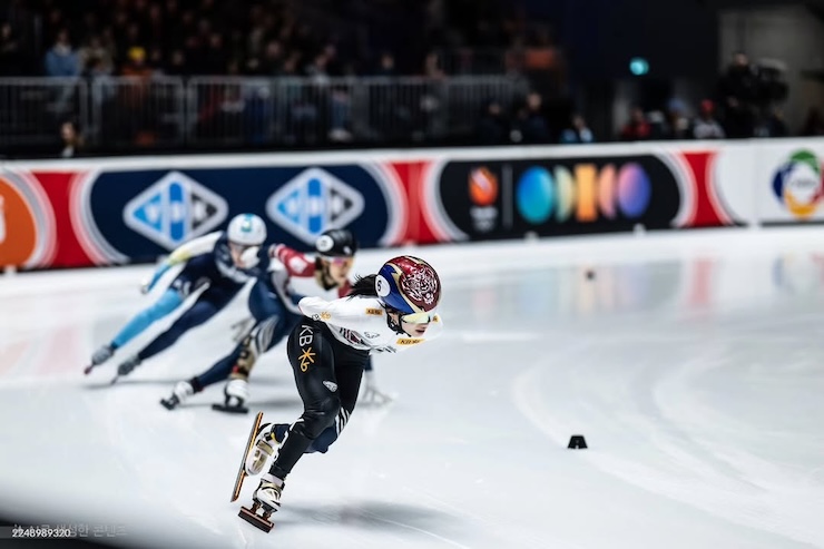 쇼트트랙 경기에서 선수들이 코너를 돌며 치열한 선두 경쟁을 벌이는 장면. 보호 장비를 착용한 선수들이 빙판 위를 빠르게 질주하고 있다.