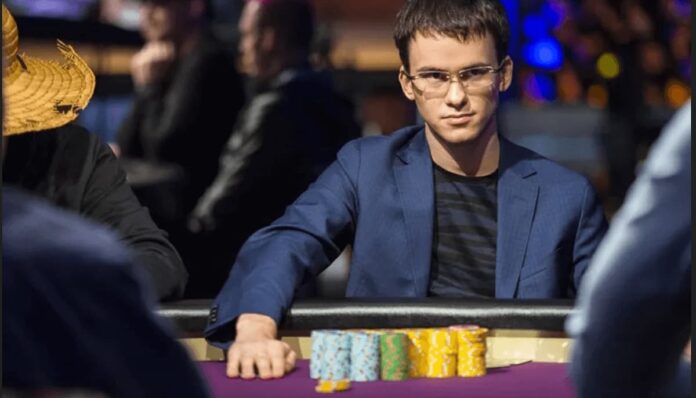 안경을 쓴 남성이 파란 재킷과 어두운 티셔츠를 입고 포커 테이블에 앉아 있으며, 앞에 여러 색상의 포커 칩이 쌓여 있다. 그는 진지한 표정으로 테이블을 바라보고 있고, 주변에는 다른 플레이어들이 흐릿하게 보인다.