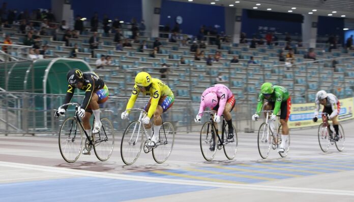 실내 벨로드롬에서 여러 명의 경륜 선수들이 선두 경쟁을 벌이며 질주하는 장면.