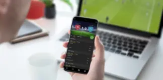 En person håller i en mobil med en bettingapp, i bakgrunden syns en laptop med en fotbollsmatch