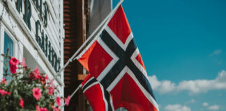 Norska flaggan vajar utomhus mot blå himmel, med blommor i förgrunden och byggnad i bakgrunden.