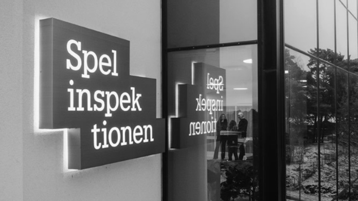 spelinspektionen-förbjuder-simba-nv Skylt med texten Spelinspektionen utanför en byggnad, speglas i fönsterglas med personer synliga inomhus.