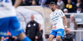 En fotbollsspelare i vit och blå matchdräkt under en match, med publiken suddig i bakgrunden.