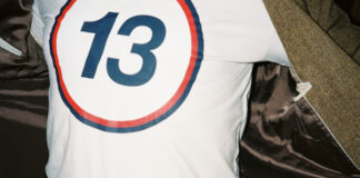 En person med öppen kavaj visar en vit tröja med en stor siffra 13 i blått inom en röd cirkel.