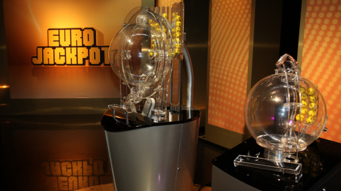 Två lottodragningmaskiner för Eurojackpot visas i en studio med bollar och logotyp i bakgrunden.