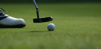 En golfare siktar på att putta en golfboll på greenen med en putter, närbild på sko och klubba.