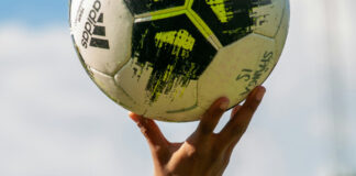 En hand håller upp en Adidas-fotboll mot himlen, klar och solig dag.