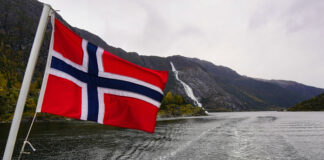 Norsk flagga vid vatten med berg och vattenfall i bakgrunden, mulen dag.