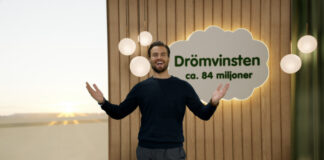 Man i mörkblå tröja står leende framför skylt med texten "Drömvinsten ca. 84 miljoner" och lampor i bakgrunden.