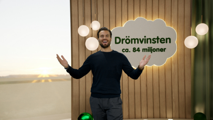 drömvinsten-till-värmland Man i mörkblå tröja står leende framför skylt med texten "Drömvinsten ca. 84 miljoner" och lampor i bakgrunden.