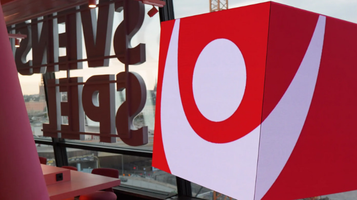 Kub med Svenska Spel-logotyp inomhus, glasvägg i bakgrunden och stadsmiljö utanför.