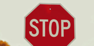 Rött stoppskylt med vit text mot ljus himmel och suddig bakgrund av träd.