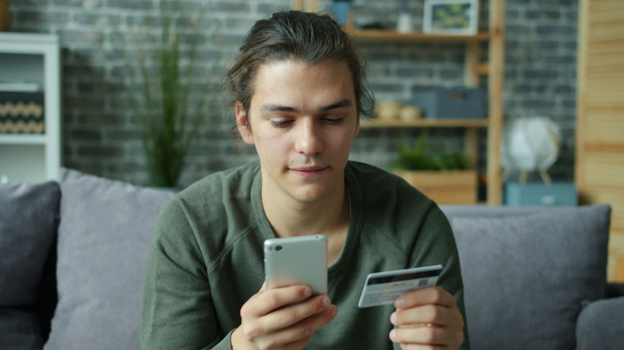 Ung man sitter i soffa och håller mobil och kreditkort när han gör ett köp online.