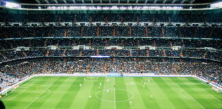 Fullsatt fotbollsstadion med pågående match sedd uppifrån, spelare spridda över den gröna planen.