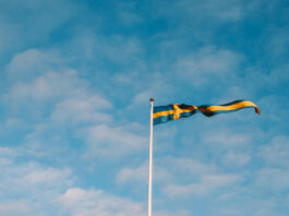 Svensk flagga som vajar i vinden mot en ljusblå himmel med lätta moln.