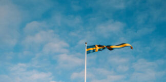 Svensk flagga som vajar i vinden mot en ljusblå himmel med lätta moln.