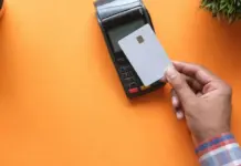 Hand håller betalkort mot kortterminal på orange bord, symbol för snabb och kontaktlös betalning.