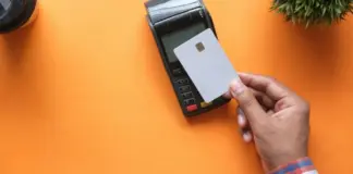 Hand håller betalkort mot kortterminal på orange bord, symbol för snabb och kontaktlös betalning.