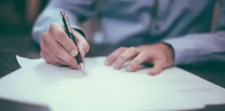 Hand som skriver under dokument med penna på skrivbord, symbol för avtal, rapportering eller juridiskt arbete.