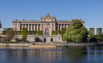 Riksdagshuset i Stockholm sett från vattnet en solig dag med grönska och klar himmel.