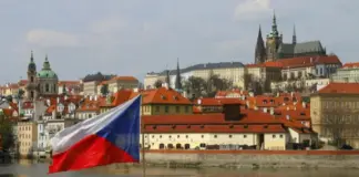 Tjeckiens flagga vid flod med stadssiluett och historiska byggnader i Prag i bakgrunden.