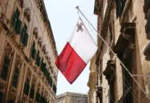 Maltas flagga hänger mellan historiska byggnader i en smal stadsgata under klar himmel