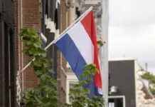 Nederländernas flagga på fasad i stadsmiljö med tegelhus och grönska längs en kanal i Nederländerna