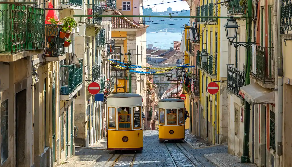 Gul spårvagn i brant gata i Lissabon med färgglada hus och utsikt över vattnet i bakgrunden