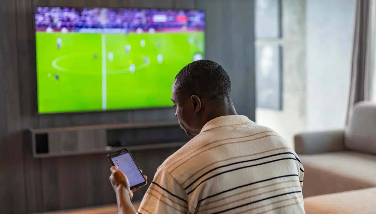 Person tittar på fotbollsmatch på tv samtidigt som mobil med bettingapp används