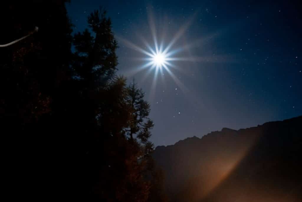 Bright starburst effect in a night sky over a forested mountain landscape, with silhouetted trees and a glowing light illuminating the horizon.