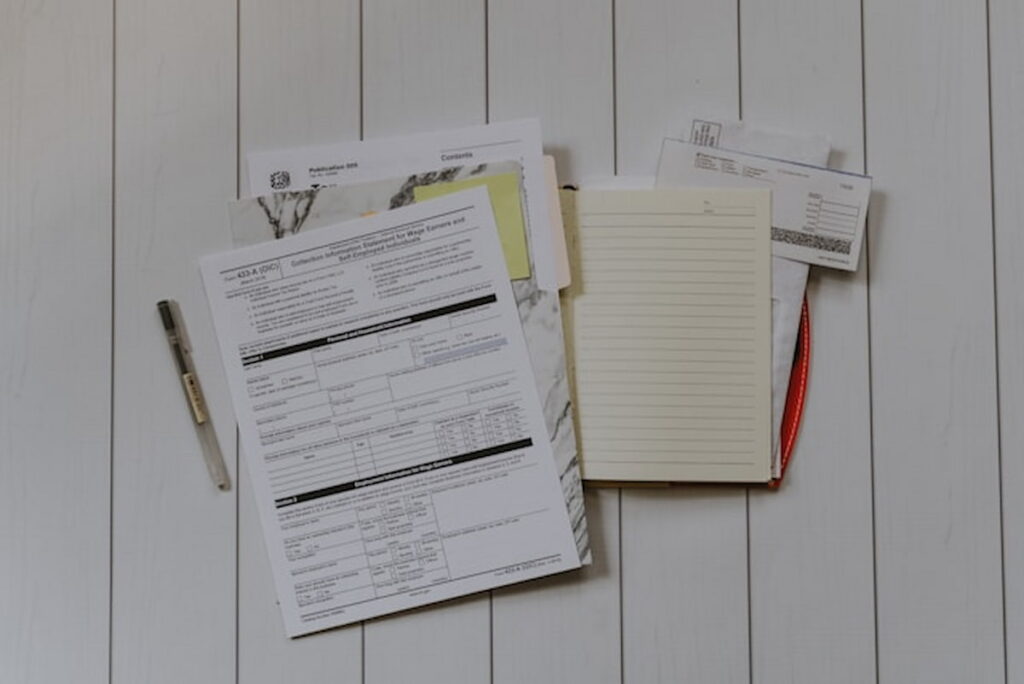 A stack of tax documents, envelopes, and a notepad lay out on a white wooden table, alongside a pen.