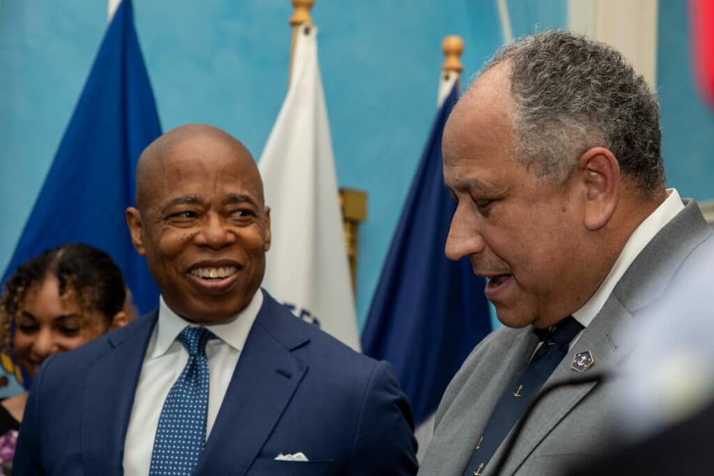 Mayor of New York City Eric Adams speaks with Secretary of the Navy Carlos Del Toro during a formal event, with flags in the background.