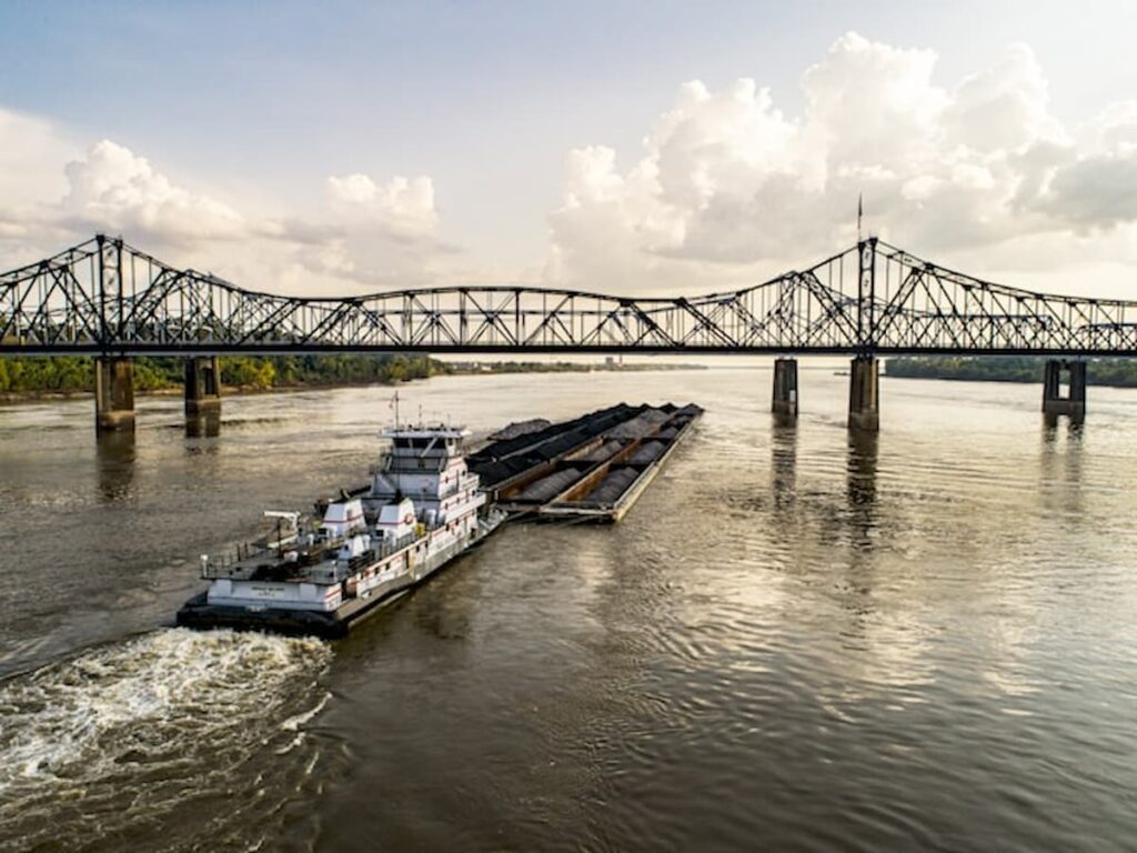 Barge traveling on the Mississippi River beneath a steel truss bridge on a partly cloudy day.