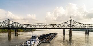 Barge traveling on the Mississippi River beneath a steel truss bridge on a partly cloudy day.