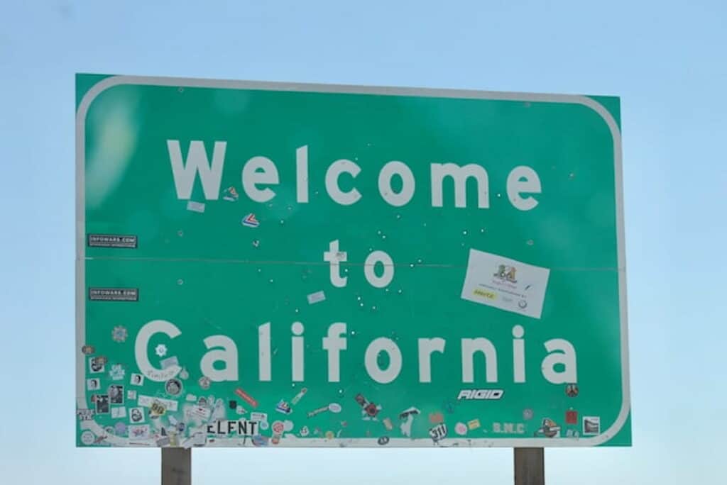 Green 'Welcome to California' road sign covered with various stickers against a clear blue sky.