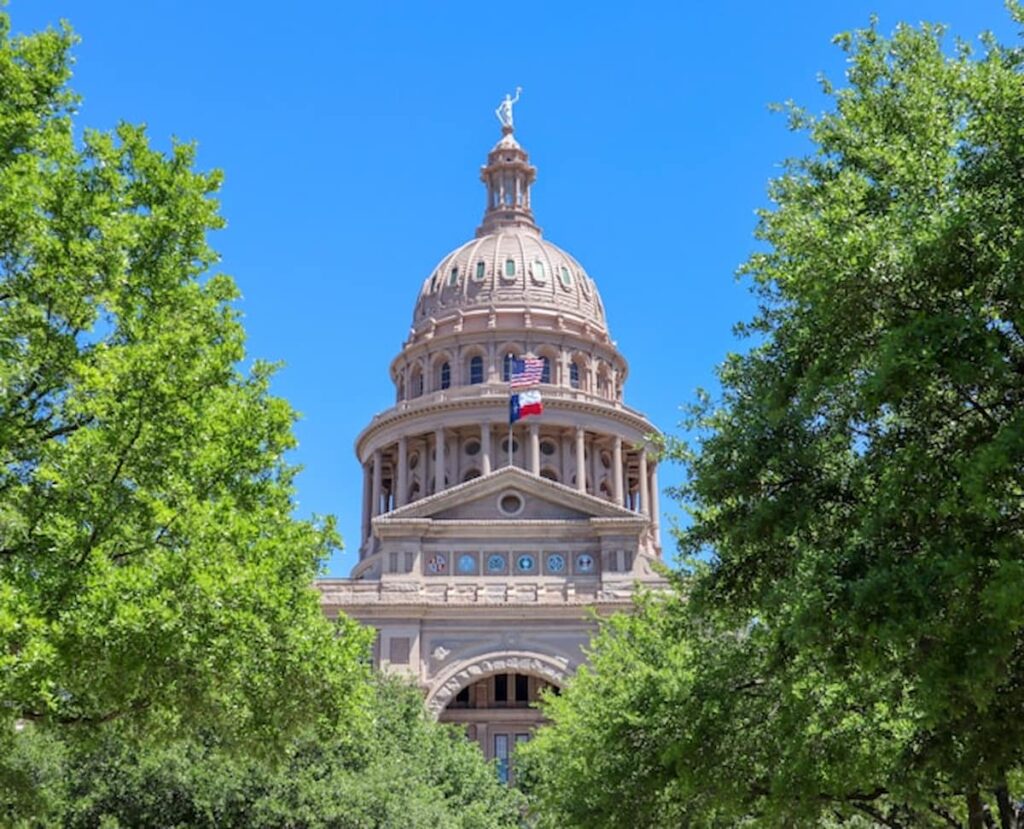 Texas State Capitol building framed by green trees under a clear blue sky.