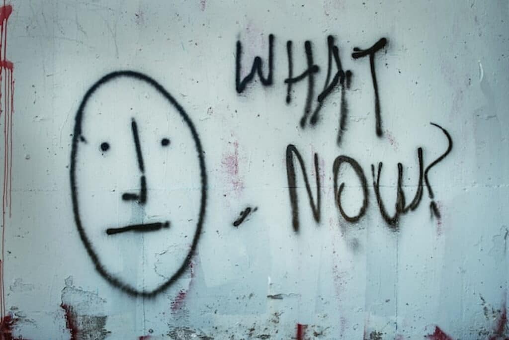 Graffiti of a simple face with the words “What Now?” on a wall