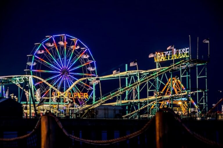 Colorful Ferris wheel and roller coaster lights glowing at night on the Atlantic City Steel Pier boardwalk.