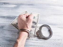 Four Arrested in Lima, Ohio for Running Illegal Gambling & Drug Operation Handcuffed hand gripping U.S. dollar bills on a wooden table
