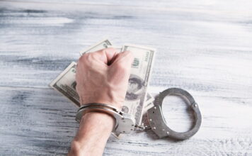 Four Arrested in Lima, Ohio for Running Illegal Gambling & Drug Operation Handcuffed hand gripping U.S. dollar bills on a wooden table