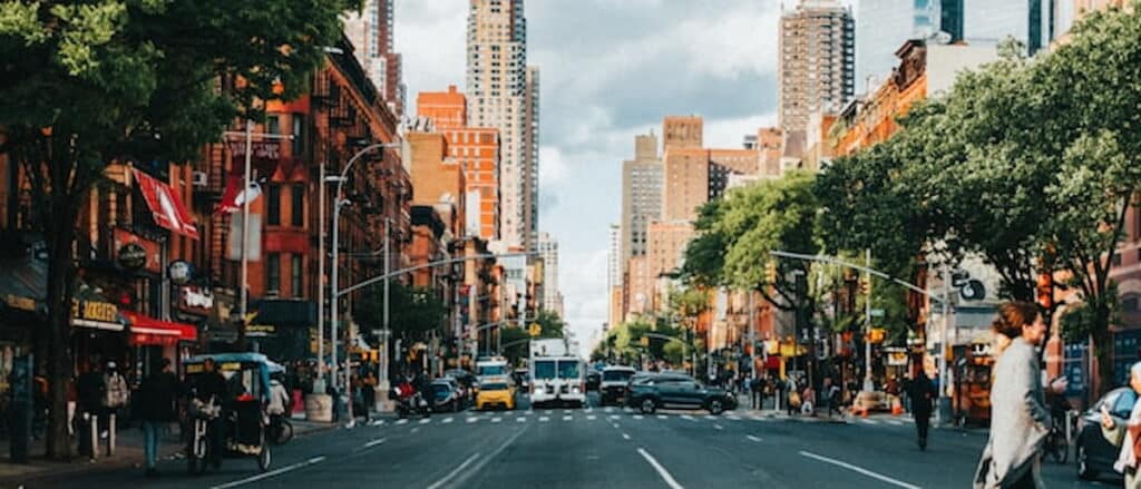 Busy street view of Hell’s Kitchen in Manhattan, with red brick buildings, trees lining the sidewalks, and traffic moving toward midtown skyscrapers.
