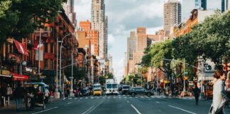 Busy street view of Hell’s Kitchen in Manhattan, with red brick buildings, trees lining the sidewalks, and traffic moving toward midtown skyscrapers.