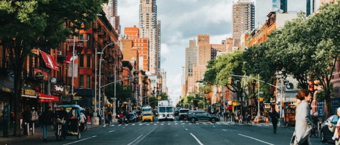 Busy street view of Hell’s Kitchen in Manhattan, with red brick buildings, trees lining the sidewalks, and traffic moving toward midtown skyscrapers.
