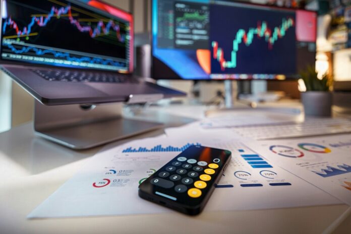 A calculator and charts sit on a desk in front of computer monitors