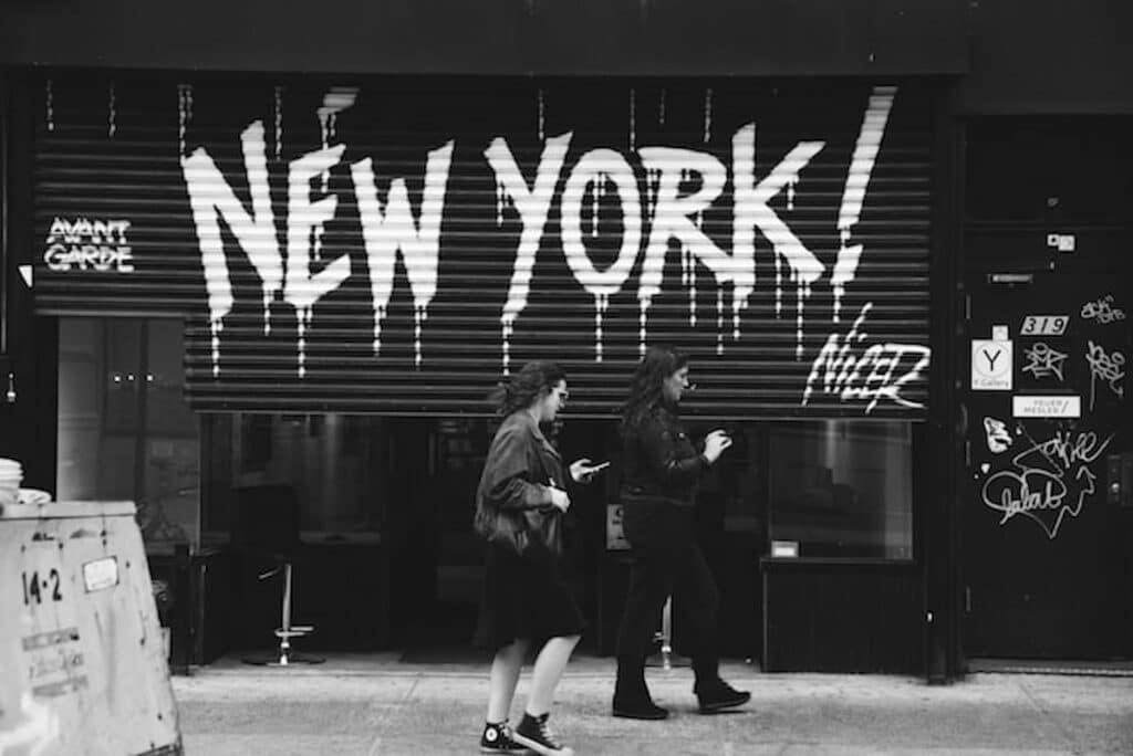 Black-and-white photo of two women walking past a storefront with bold graffiti reading “NEW YORK!” on its metal shutter.