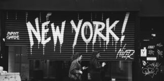 Black-and-white photo of two women walking past a storefront with bold graffiti reading “NEW YORK!” on its metal shutter.