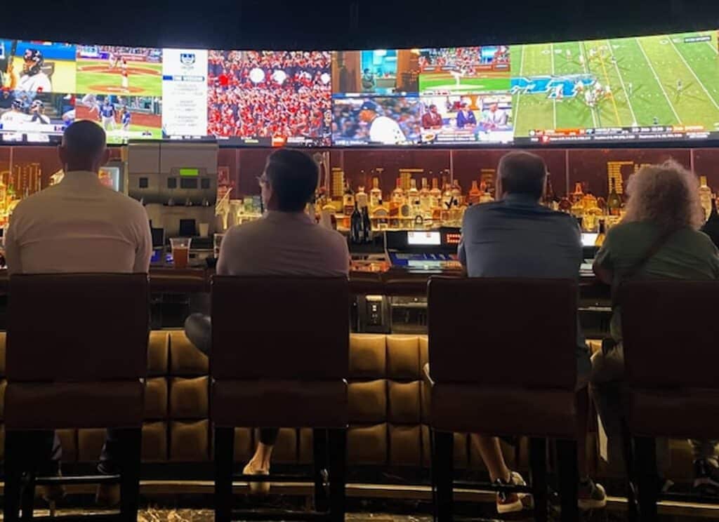 People sitting at a bar watching multiple live sports broadcasts on large screens above a row of liquor bottles.