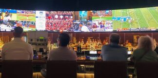 FanDuel Partner CME Moves Into Sports Prediction Markets People sitting at a bar watching multiple live sports broadcasts on large screens above a row of liquor bottles.